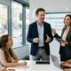 Une photographie de style professionnel montrant un homme et une femme coanimant une réunion d'équipe. Ils se tiennent debout devant un écran affichant « Q3 STRATEGY AND GROWTH » et un tableau blanc rempli de graphiques. Trois collègues assis à une table blanche les écoutent attentivement dans un bureau moderne et lumineux avec de grandes baies vitrées.